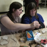 A woman helping a person with disabilities paint clay ornaments.