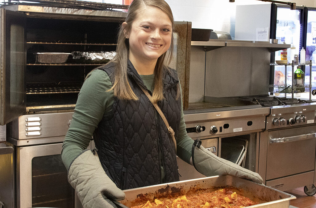A young woman in a commercial kitchen preparing a large pan of baked pasta.