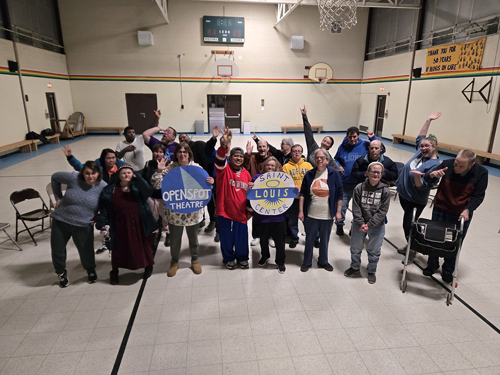 A group of adults dressed in casual clothes standing in silly poses in the St. Louis Center gym.